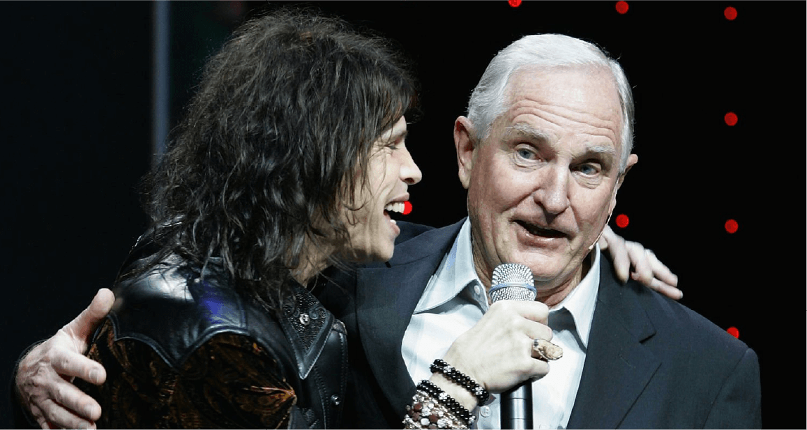 Steven Tyler and Craig Barrett sing into a microphone together on stage.